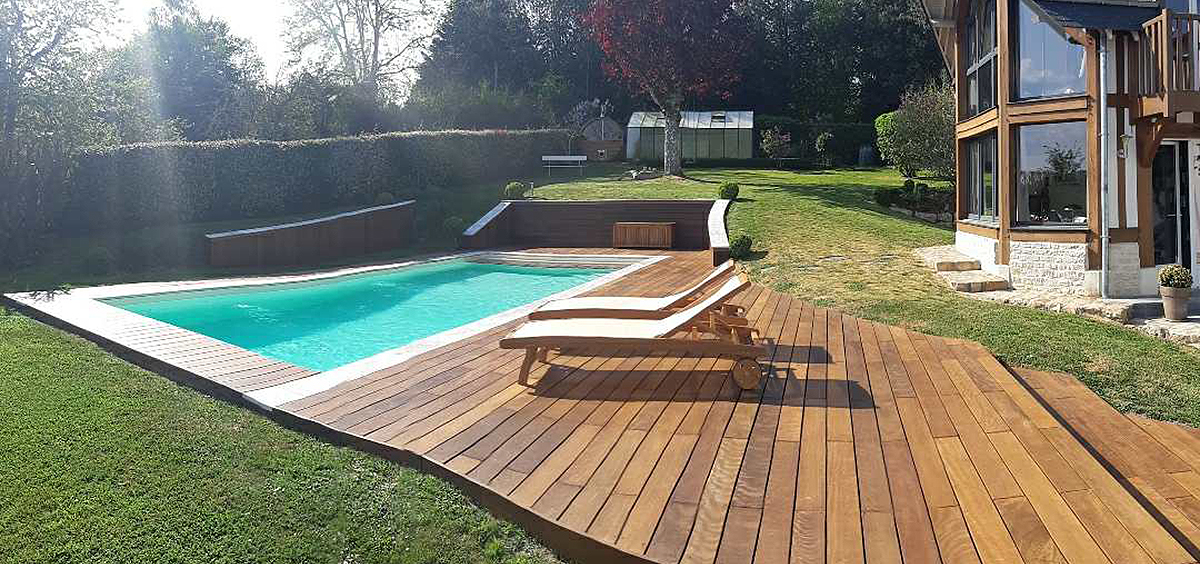 Terrasse en bois exotique brun doré entourant une piscine turquoise dans un jardin paysager, avec transats en bois et maison contemporaine à ossature bois en arrière-plan, illustrant un aménagement extérieur haut de gamme et chaleureux.