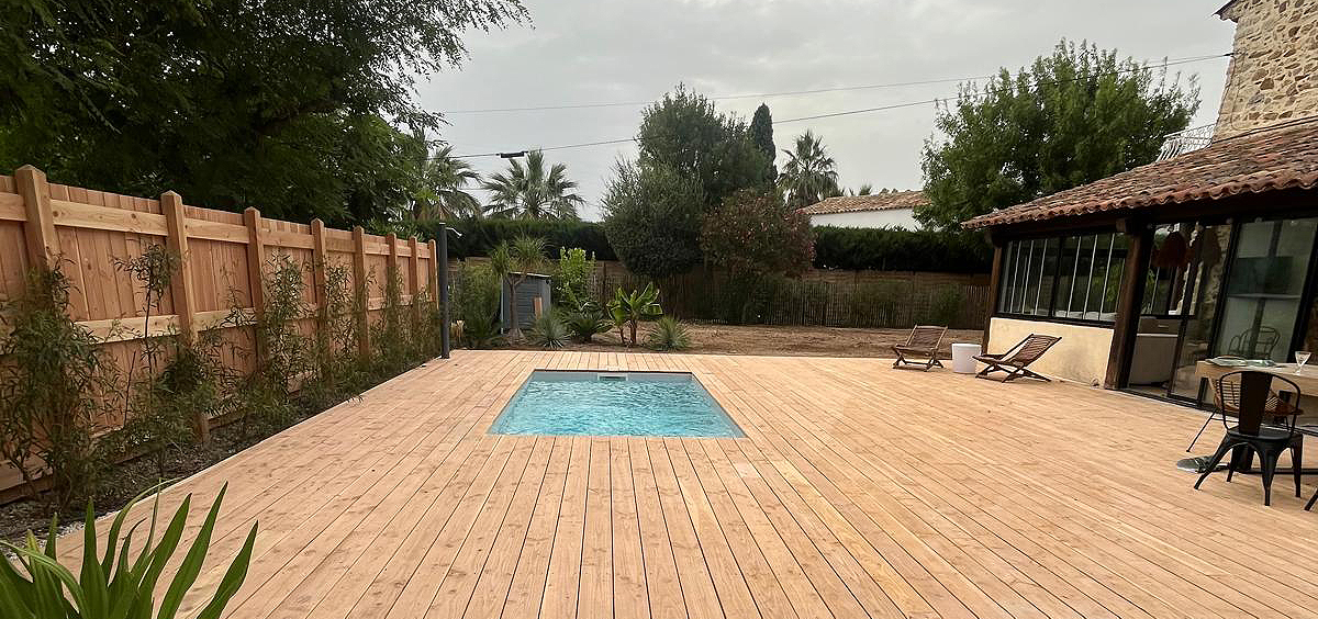 Terrasse extérieure en bois clair entourant une petite piscine rectangulaire, adossée à une maison en pierre avec toiture en tuiles, dans un jardin verdoyant avec clôture bois et végétation méditerranéenne, illustrant un aménagement naturel et chaleureux.