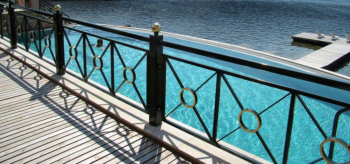 Terrasse en bois avec garde‑corps métallique décoratif surplombant une piscine et un lac, ambiance estivale.