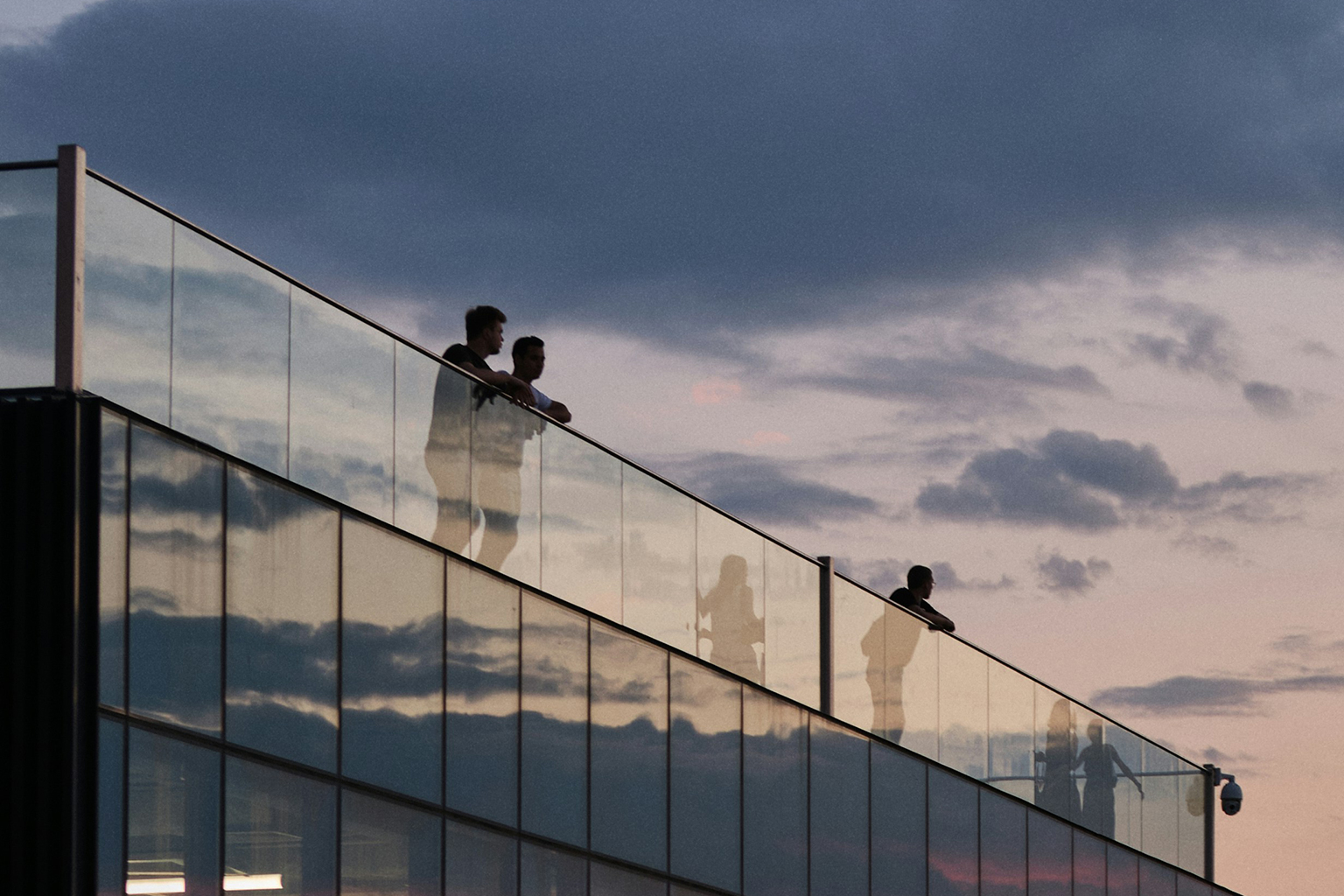Terrasse en bois avec garde‑corps en verre transparent sur bâtiment moderne, au coucher du soleil.