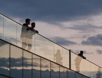 Terrasse en bois avec garde‑corps en verre transparent sur bâtiment moderne, au coucher du soleil.