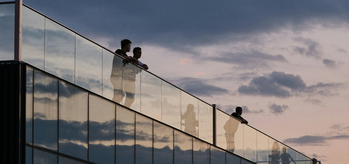 Terrasse en bois avec garde‑corps en verre transparent sur bâtiment moderne, au coucher du soleil.