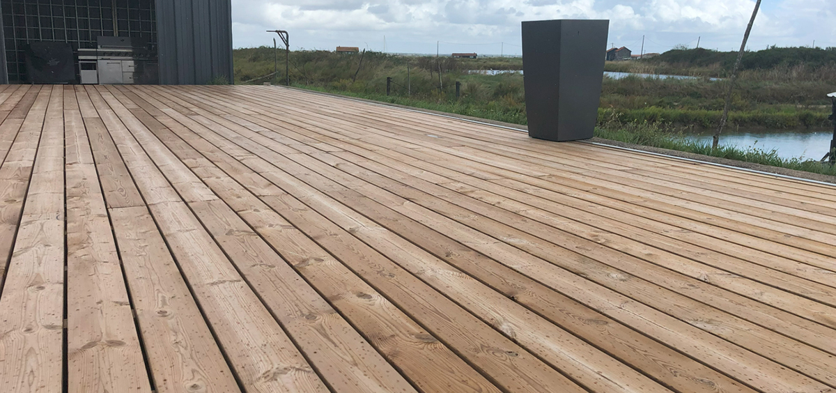 Grande terrasse extérieure en bois Pin clair, installée au bord d’un plan d’eau, avec une structure moderne en métal et un espace barbecue, illustrant la beauté naturelle et la durabilité du bois pour les aménagements extérieurs.