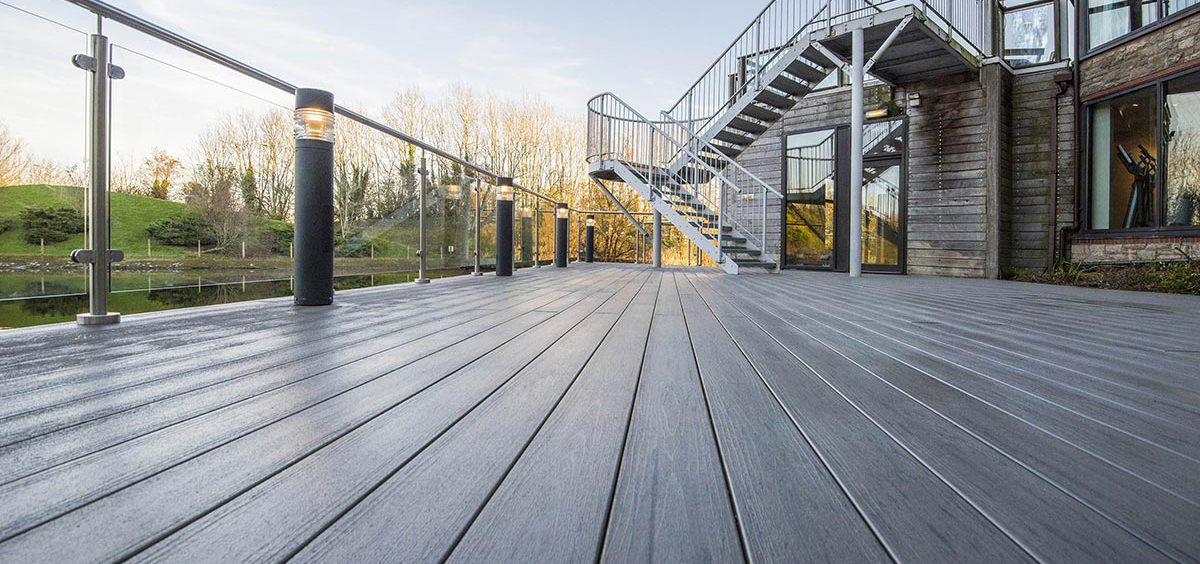 Terrasse extérieure en lame composite grise avec garde-corps en verre et poteaux en acier, éclairée par des lampes cylindriques, adossée à une maison contemporaine en bois et métal, offrant une vue dégagée sur un jardin et un plan d’eau.